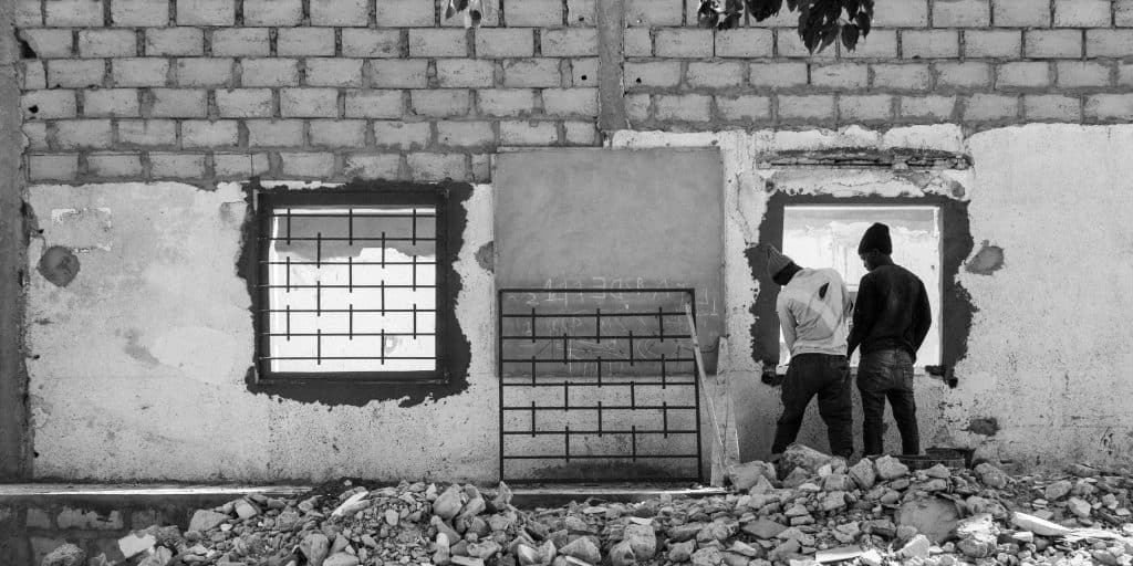 Black and white photo. A wall with two empty square holes where windows will be inserted. One has a large metal grate over it. The second grate leans up against the wall while two men prepare to mount it over the second window.