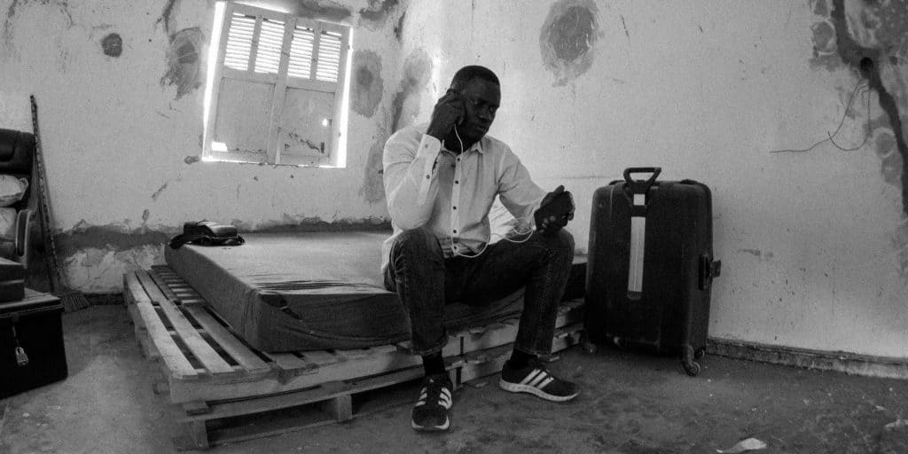 Black and white photo: A large Senegalese man wearing jeans, Adidas running shoes, and a nicely pressed white button-down shirt sits on the edge of a mattress. The mattress is on top of a short stack of wooden pallets. The house is under construction, so the walls are pitted and peeling with cement scars. The man holds a phone to his ear while a cable connects it to the mobile charger in his other hand.