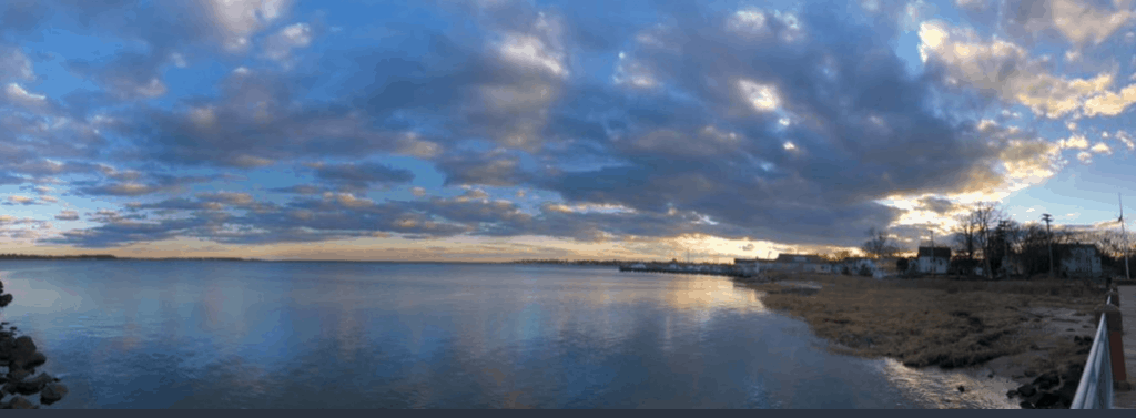 early dusk on Long Island Sound with clouds spread across a blue sky and the sun setting in the lower right hand corner of the image