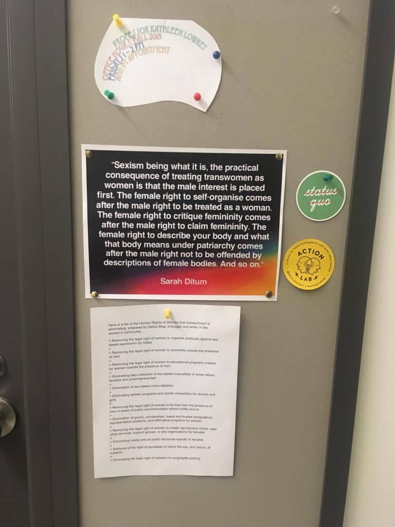 Photo of office door which includes a small cutout with contact information, two small circular stickers, and 8.5x11 papers with gender critical commentary