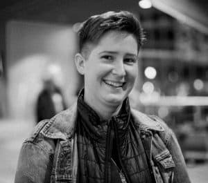 The photo is of Rine Vieth, in black and white. They have short hair, are smiling slightly, and are wearing a denim jacket over a puffer jacket.