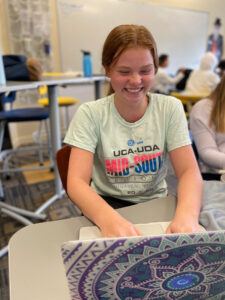 A high school student, the author, is a young woman with red hair and big grin, sits typing at her laptop.